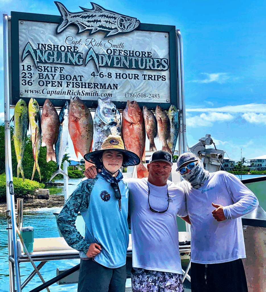 Three fishermen posing with a variety of freshly caught fish, including Yellowtail Snapper, in front of a sign for Capt. Rich Smith's Angling Adventures, promoting fishing charters in the Florida Keys.