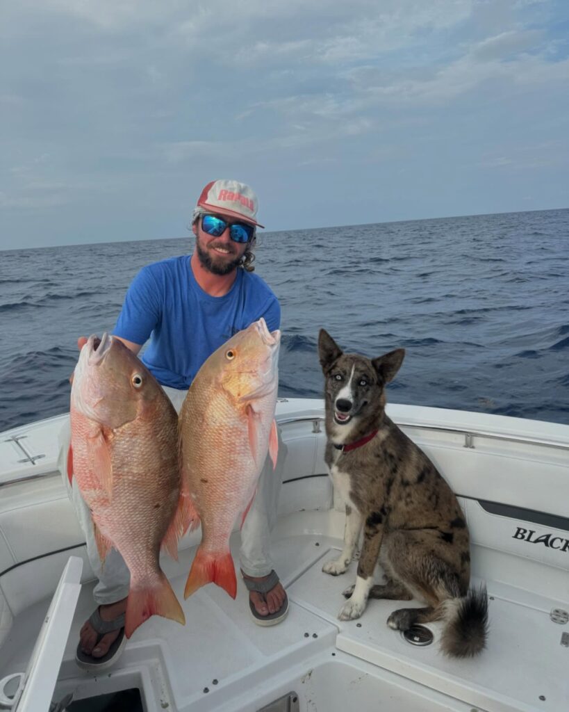 Man holding two large fish on a fishing boat, with a dog sitting beside him, showcasing a successful fishing trip in the Florida Keys.