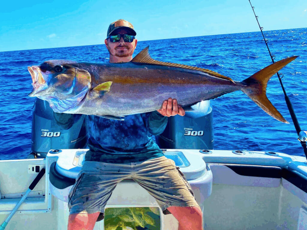 Man holding a large amberjack while fishing in the Florida Keys, with blue ocean waters and boat motors in the background, showcasing the thrill of wreck fishing.