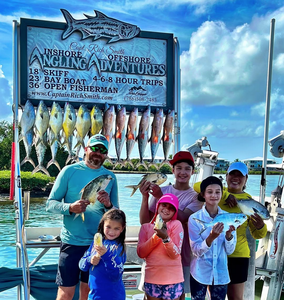 Family fishing charter group displaying their catch, including various fish species, in front of a fishing boat, with a sign promoting Angling Adventures in the Florida Keys.