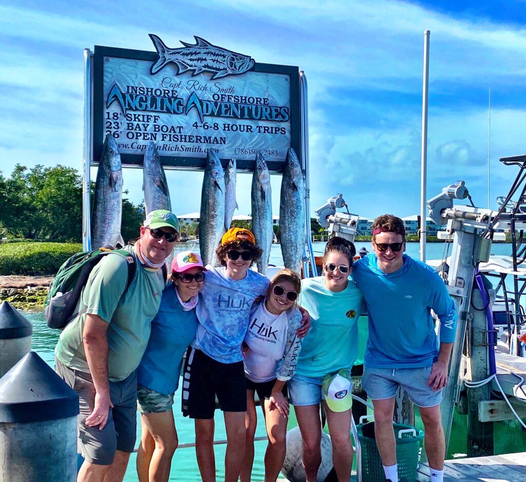 Group of anglers celebrating their catch of giant Kingfish at a fishing charter dock in the Florida Keys, with a sign for Capt. Rich Smith's Angling Adventures in the background, showcasing a family-friendly fishing experience.