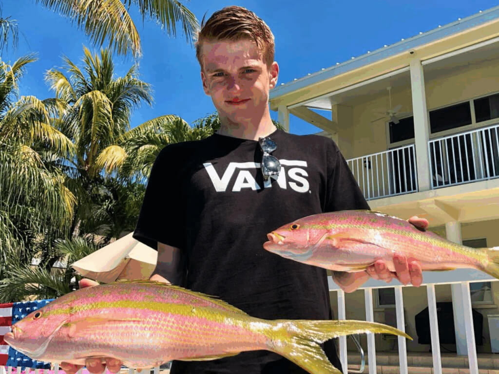Young man holding two Yellowtail Snapper, showcasing a successful reef fishing catch in the Florida Keys, with palm trees and a house in the background.